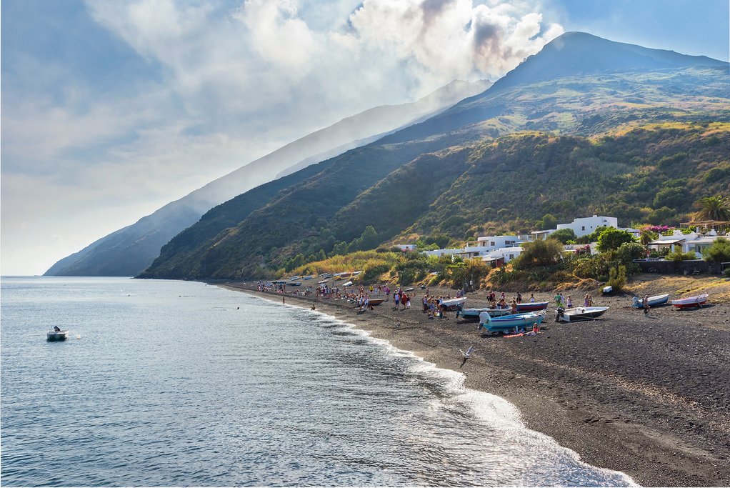 Italy - Sicily - Stromboli - People relax on the black sand beach with Stromboli in background