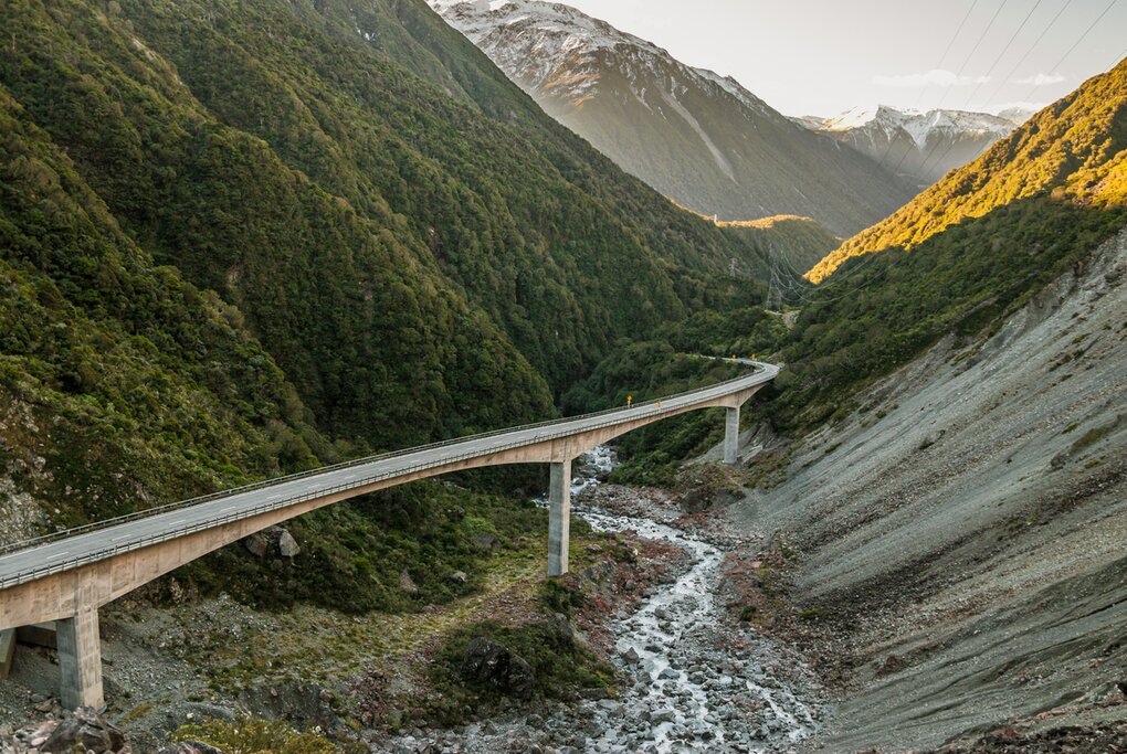 an elevated road crosses between two mountain slopes with a shallow river below in Arthur's Pass, New Zealand