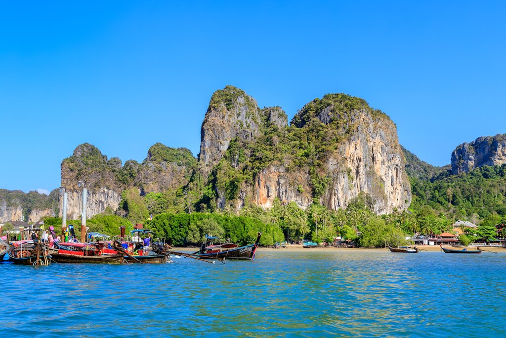 Turquoise crystal clear sea water with limestone cliff and mountain at Railay East Beach with several long tail boats moored offshore.