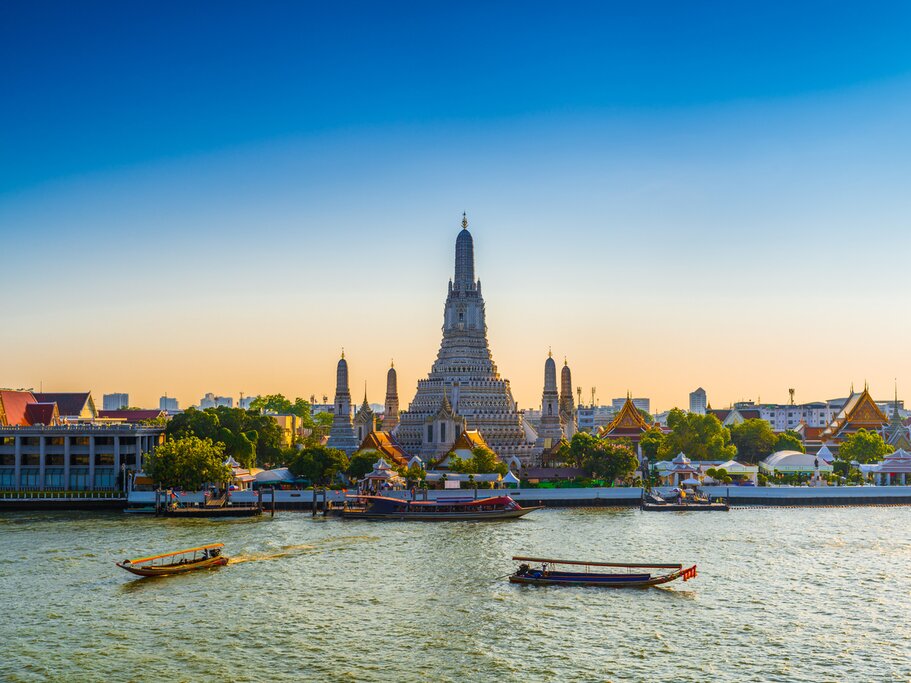 Photo across the water of Wat Arun (Temple of Dawn) a sunset with a yellow-blue sky. A couple of long tail boats float by in the foreground.