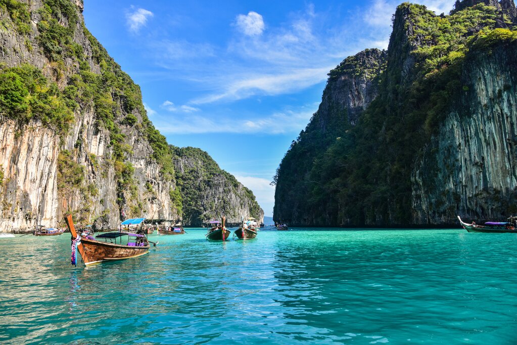 Photo of Pileh Lagoon in the Phi Phi Islands of Thailand. The camera faces several long tail boats head-on as they pass through calm turquoise water surrounded by limestone cliffs
