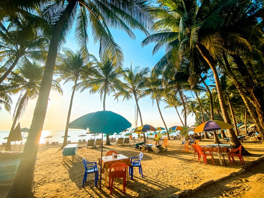 Photo of Surin Beach in Phuket, with palm trees, colorful plastic tables and chairs, and umbrellas