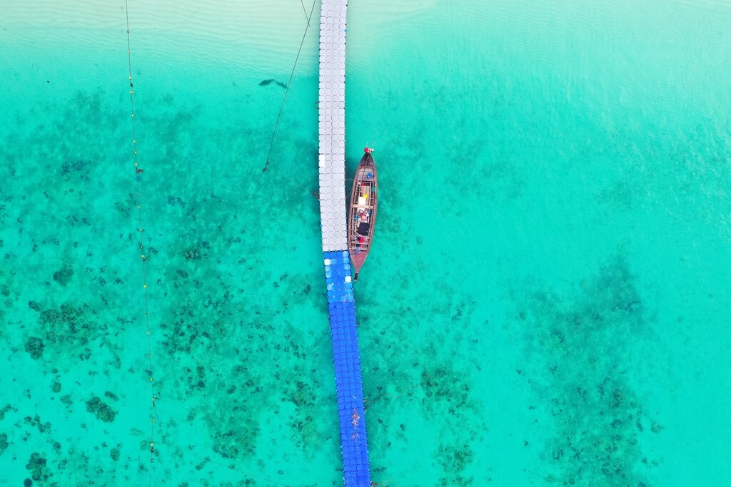 Birds-eye view of a long thin pier over shallow, clear tropical water with a small boat docked alongside.