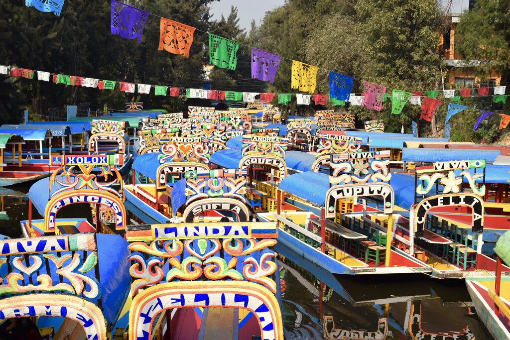 A collection of trajineras (colorful wooden boats) sit empty in a canal in Zochimilco with strings of colorful paper cut outs hanging about them