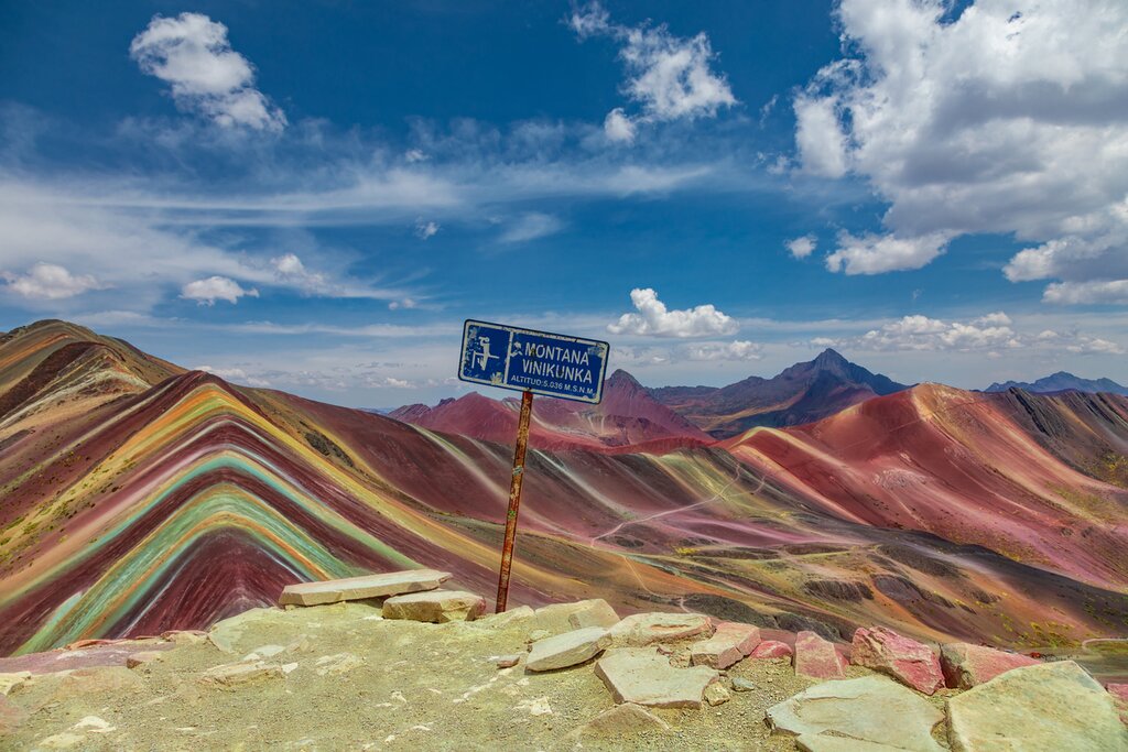 View of Vinicunca, "Rainbow Mountain," from the summit of a neighboring slope, with a blue sign reading "Montana Vinikunka Altitud: 5.036 M.S.N.M."