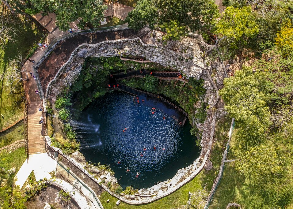 Aerial view of a cenote in Mexico. Wooden walkways surround a round hole in the ground filled with turquoise water with people swimming