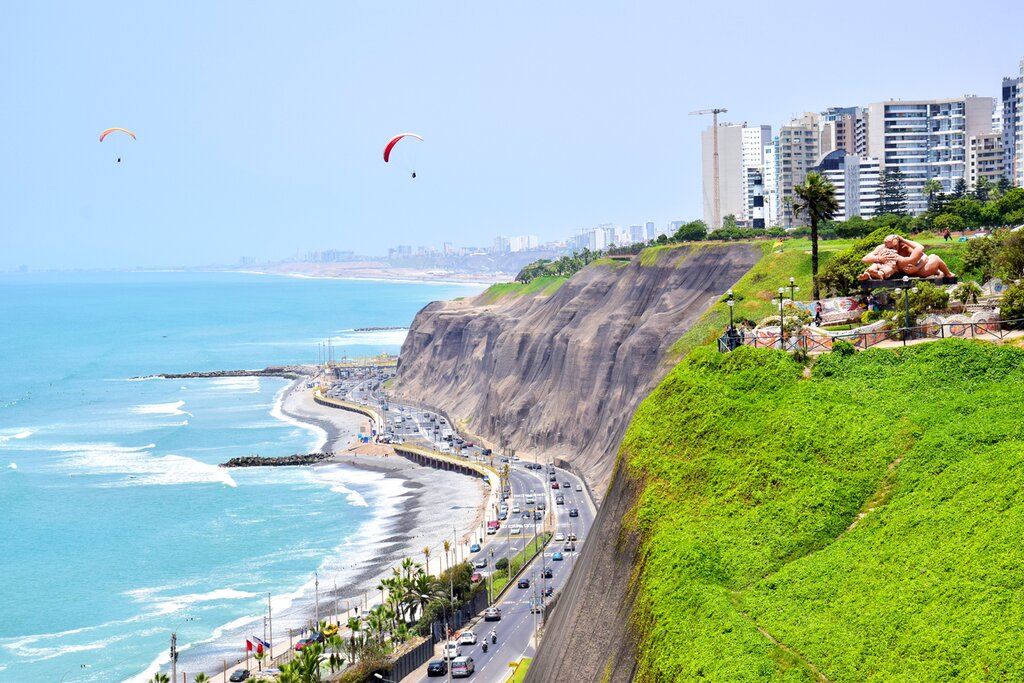 Aerial view of the coast of Lima, with the road and beach on the left and cliffs on the right. Two paragliders soar above the scene.