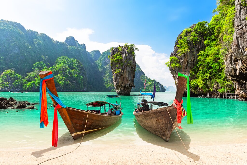 Photo from the beach of James Bond Island, a tall, thin limestone protrusion in a bright aqua sea. Two wooden long tail boats sit on the beach in the foreground, facing the camera