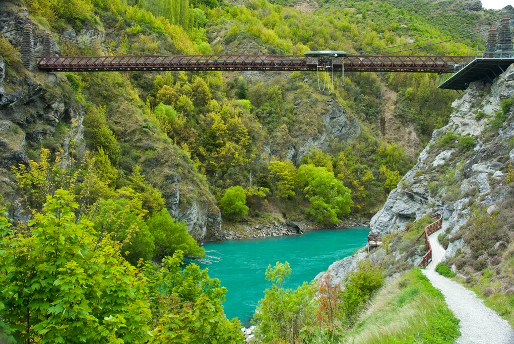 Kawarau Bridge in Queenstown, suspended over turquoise water with bright green foliage in the background. Bungee jumping ropes can be seen attached to the bridge.