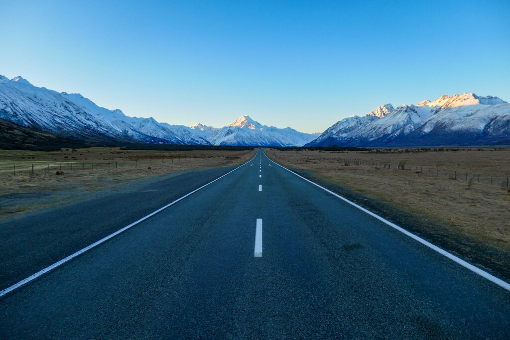 The road to Aoraki Mount Cook. The road takes up the center of the photo and leads straight ahead to white mountains in the background