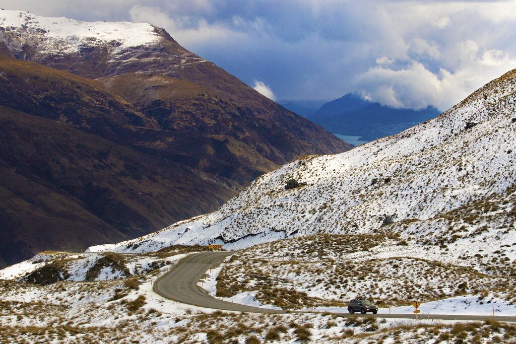 Crown Range Road with a jeep in the foreground and snow capped mountains in the background on a partly cloudy day